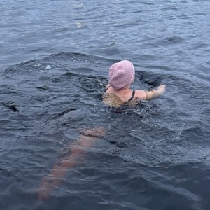 Henkilö, jolla on vaaleanpunainen uimalakki, ui suuressa, rauhallisessa vesistössä lähellä kallioista rantaviivaa, ja taustalla näkyy rakennuksia ja puita pilvisen taivaan alla.