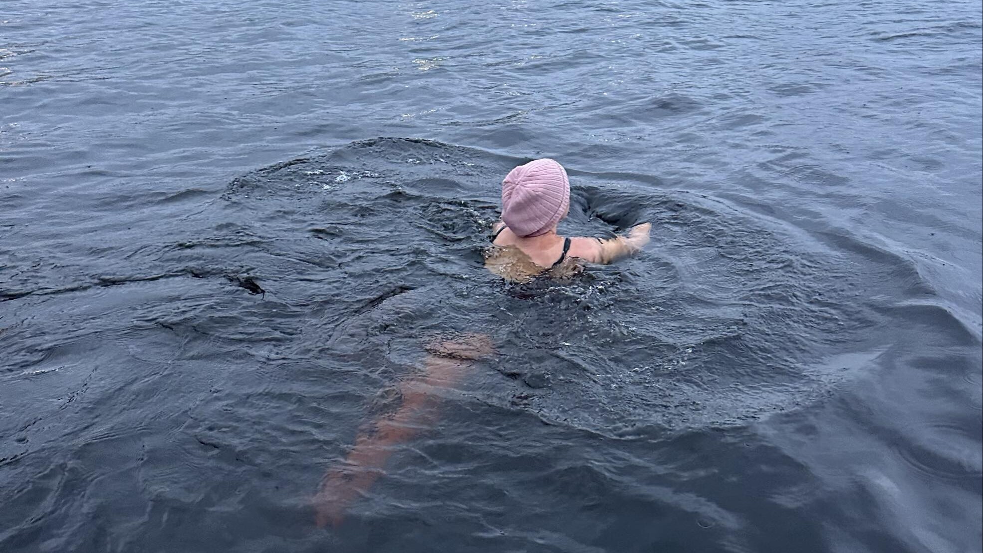 Henkilö, jolla on vaaleanpunainen uimalakki, ui suuressa, rauhallisessa vesistössä lähellä kallioista rantaviivaa, ja taustalla näkyy rakennuksia ja puita pilvisen taivaan alla.