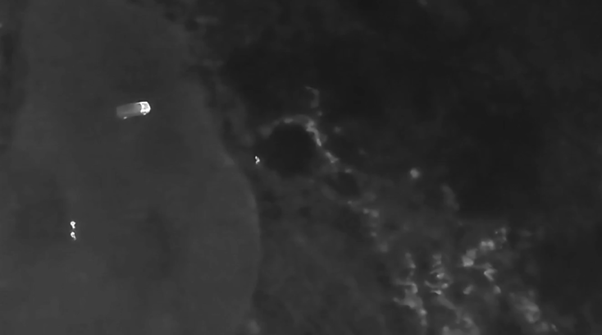 Mustavalkoisessa ilmakuvassa tai lämpökuvassa näkyy vene vedessä lähellä rantaviivaa, ja yksi henkilö on vedessä lähellä rantaa ja toinen lähellä venettä. Ympäröivä vesi ja maa näyttävät tummilta, ja niissä on joitakin vaaleita kohtia.