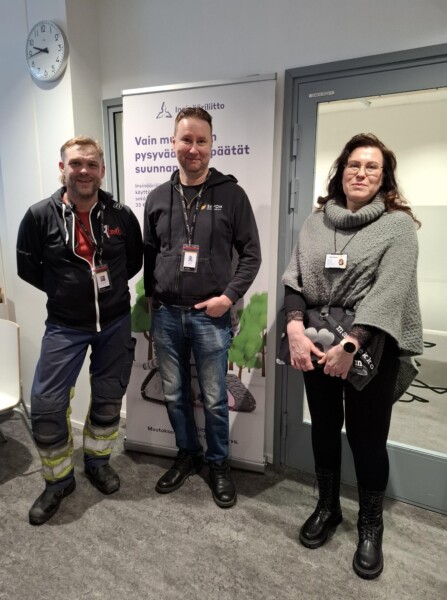 Kolme ihmistä seisoo sisätiloissa banderollin ja suljetun oven edessä, ja kaikilla on henkilötunnukset. Yhdellä henkilöllä on työvaatteet, toisella vapaa-ajan vaatteet ja kolmannella poncho, jossa on vesipullo kädessä. Seinällä näkyy kello.