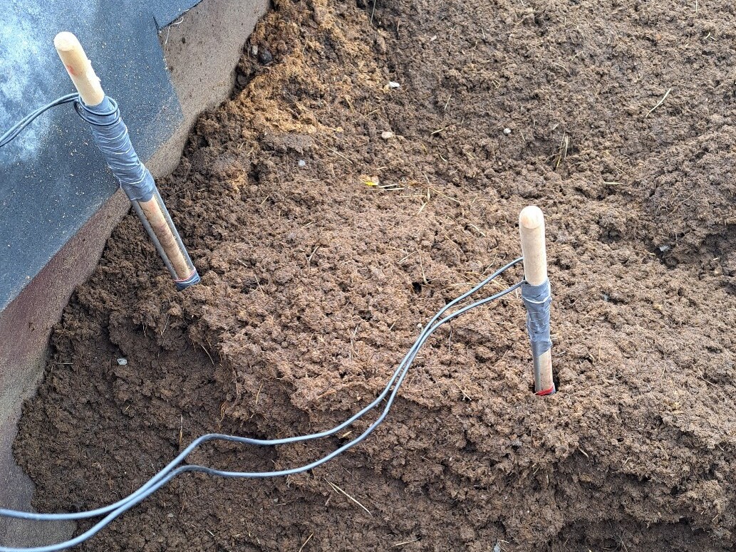 Kaksi puukahvaista työkalua, joiden ympärille on kiedottu naruja, työnnetään suureen multa- tai kompostikasaan, joka on mustilla ja punaisilla sivuilla varustetun astian sisällä.