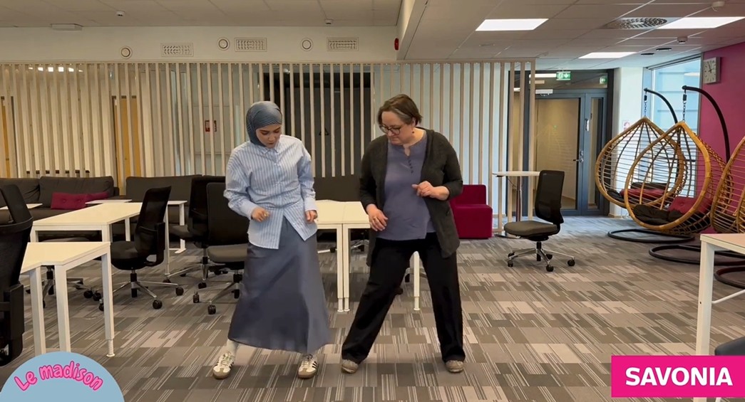 Kaksi naista tanssii yhdessä avoimessa, modernissa toimistotilassa, jossa on pöytiä, tuoleja ja riipputuolia. Toisella on hijab ja hame, toisella silmälasit ja tummat housut. Le madison- ja SAVONIA-logot ovat näkyvissä.