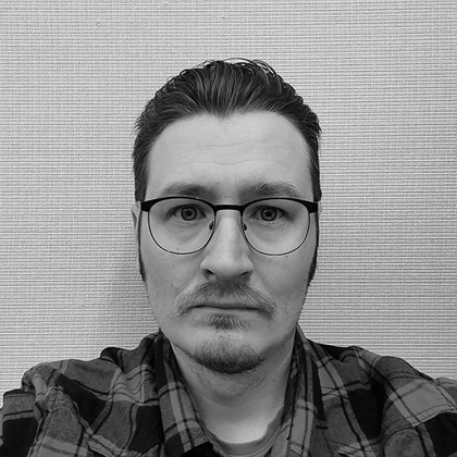 Mustavalkoinen valokuva henkilöstä, jolla on lyhyet tummat hiukset ja silmälasit, päällään ruutupaita, katsoo suoraan kameraan neutraali ilme. Taustalla on kuvioitu, vaalea seinä.