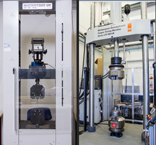 Kaksi erilaista mekaanista testauskonetta laboratoriossa; molemmat ovat korkeita, pystysuoria laitteita, joissa on metallirunko ja erilaisia antureita ja joita käytetään materiaalien testaamiseen jännityksen tai puristuksen alaisena.