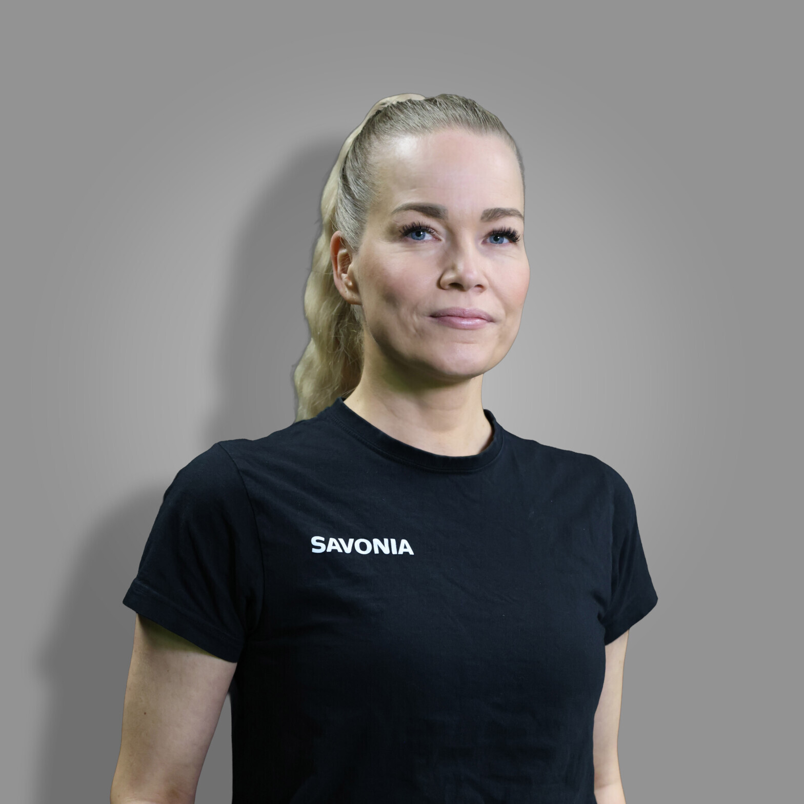 Nainen, jolla on vaaleat hiukset poninhännässä ja päällään musta t-paita, jossa lukee SAVONIA, seisoo harmaata taustaa vasten ja katsoo hieman sivulle.