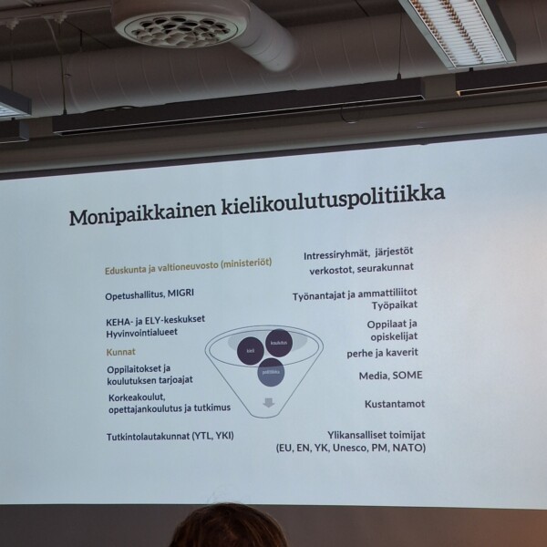 Suomenkielisessä diassa Monipaikkainen kielikoulutuspolitiikka esitetään kielikoulutuspolitiikan sidosryhmiä, kuten eduskunta, ministeriöt, koulut, kunnat, työntekijät, tiedotusvälineet ja kansainväliset järjestöt, keskeisellä kaaviolla.