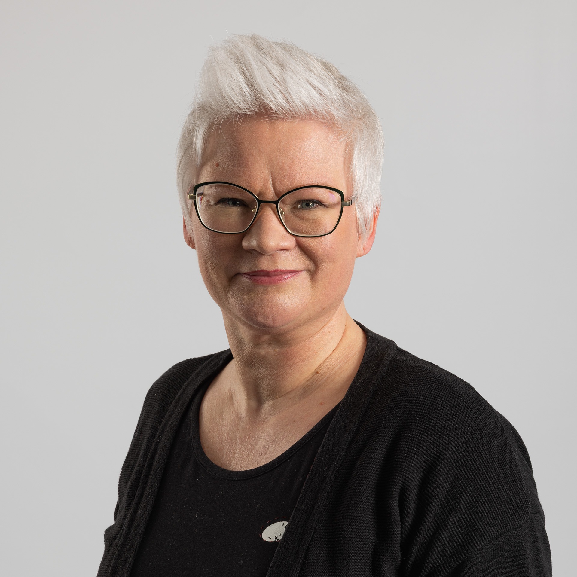 Lyhyet, valkoiset hiukset ja silmälasit omaava henkilö hymyilee kameralle, yllään musta toppi ja villatakki vaaleanharmaalla taustalla.