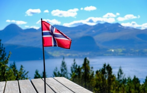 Pieni Norjan lippu kepissä seisoo puupöydällä, ja taustalla on maisemallinen näkymä järvelle, vihreille puille ja vuorille kirkkaan sinisen taivaan alla.