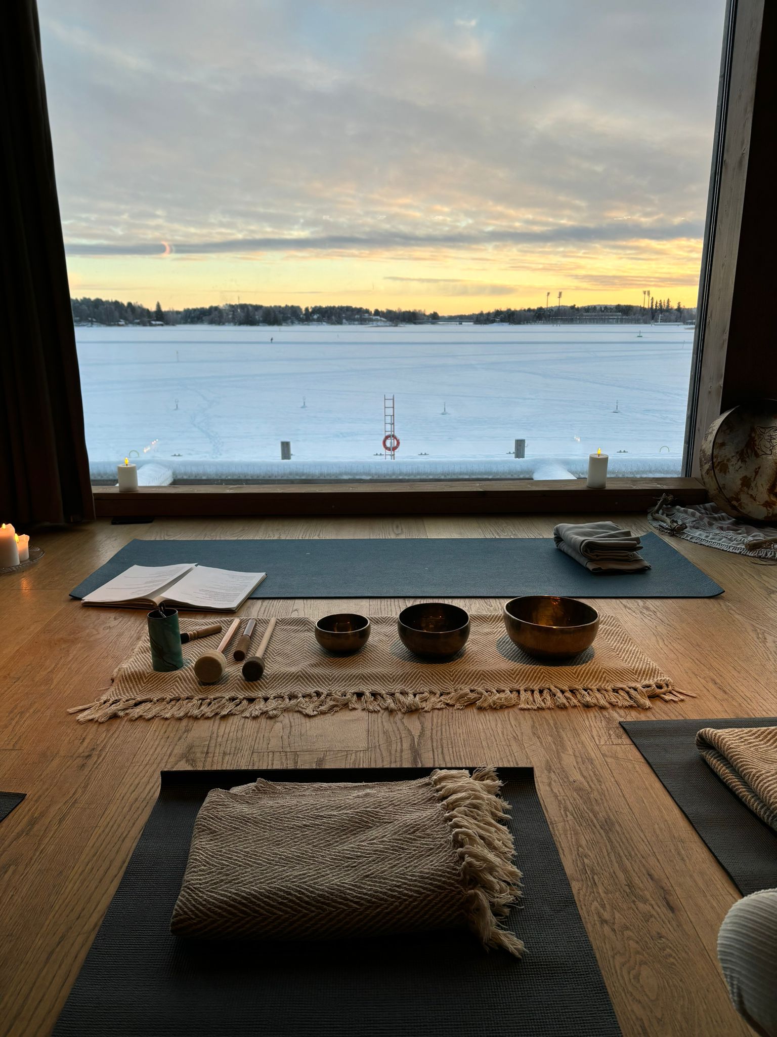 Seesteinen sisäjooga-asetelma on vastapäätä suurta ikkunaa, josta avautuu näkymä lumiselle maisemalle auringonlaskun aikaan. Puulattialla on joogamattoja, peittoja ja laulukuppeja, ja kynttilät luovat lämpimän ja rauhoittavan tunnelman.