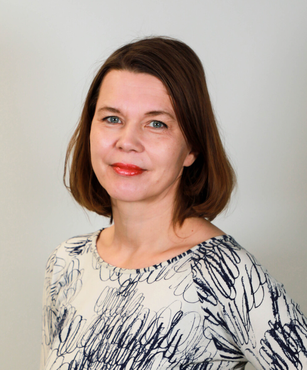 Nainen, jolla on suorat ruskeat hiukset ja vaalea iho, seisoo tavallista taustaa vasten, yllään valkoinen toppi, jossa on sinisiä abstrakteja kuvioita. Hän katsoo kameraan ja hymyilee lempeästi.