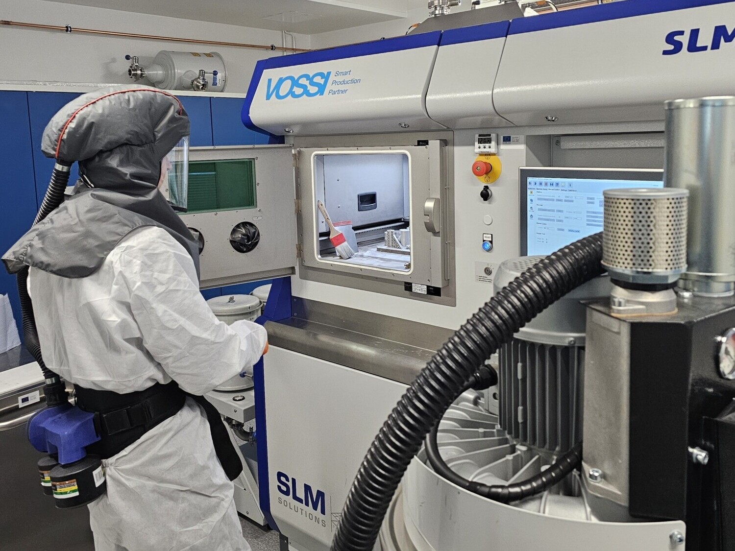 Suojavarusteisiin pukeutunut henkilö käyttää 3D-metallitulostinta laboratorioympäristössä, valvoo prosessia ikkunan kautta ja käyttää koneen ohjauspaneelia, jossa on merkintä "SLM 280".".