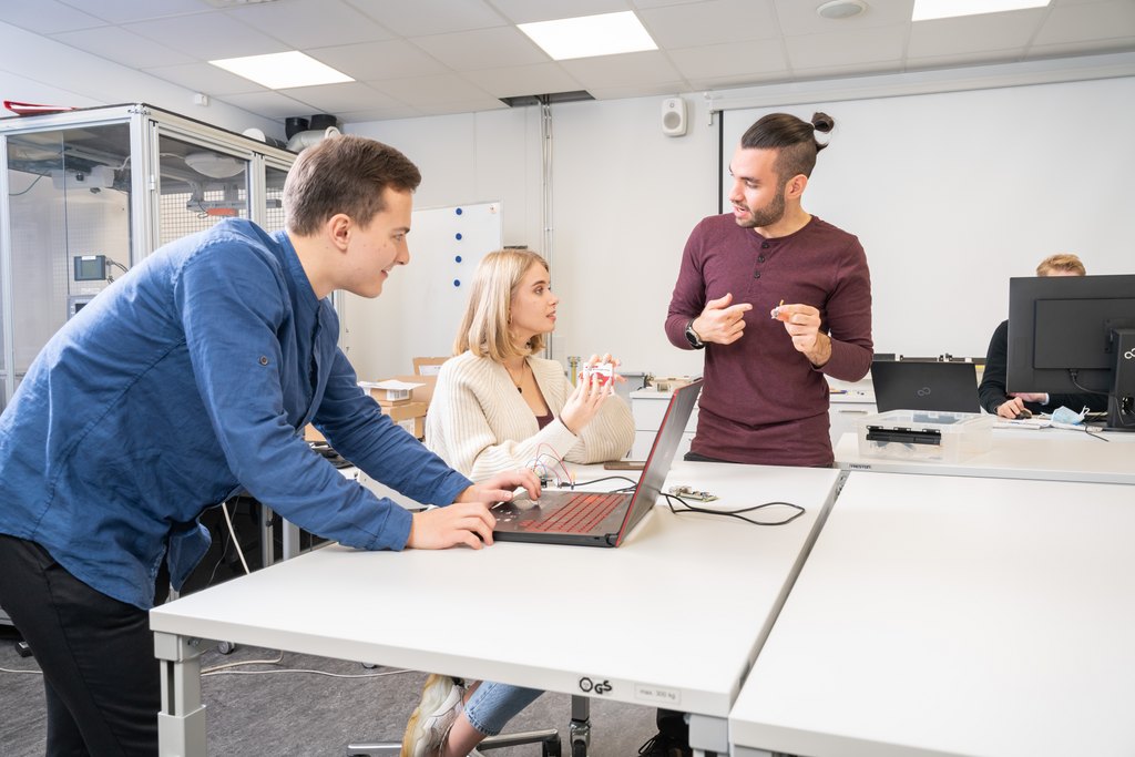 Kolme nuorta aikuista tekee yhteistyötä valoisassa toimistossa tai luokkahuoneessa. Yksi seisoo ja käyttää kannettavaa tietokonetta, toinen istuu pitelemässä elektronista laitetta, ja kolmas seisoo ja elehtii puhuessaan. Heidän ympärillään on pöytiä, tietokoneita ja toimistolaitteita.