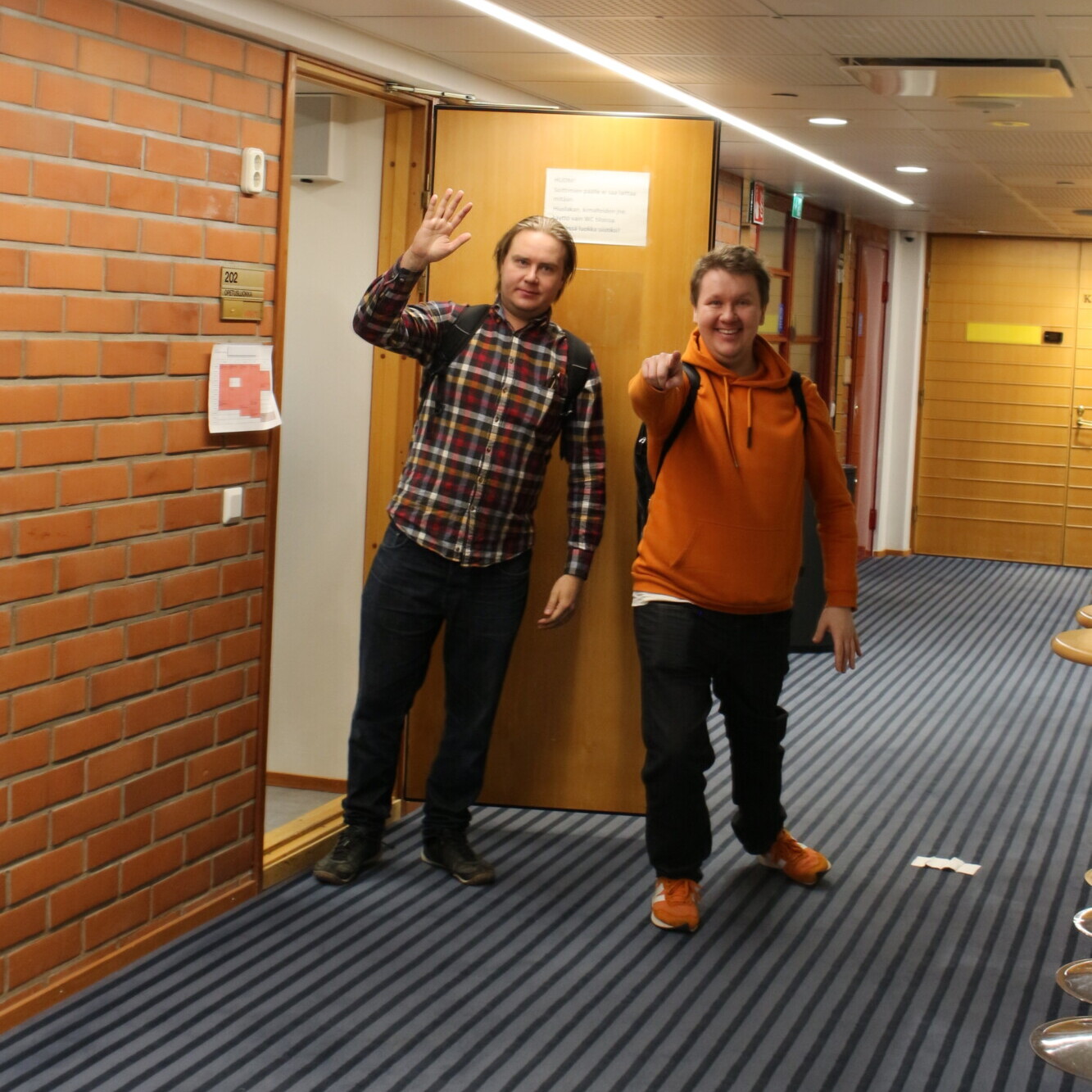 Kaksi miestä seisoo käytävällä hymyillen ja elehtien kohti kameraa. Toisella on ruudullinen paita ja farkut, toisella oranssi huppari ja kengät. Käytävässä on raidallinen matto ja tiiliseinät.