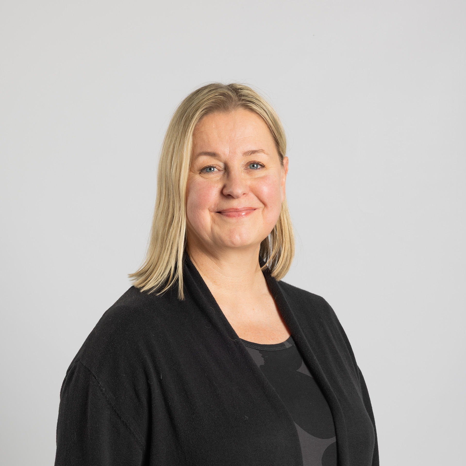 Vaaleatukkainen nainen, jolla on musta villatakki ja tumma toppi, hymyilee lempeästi seisoessaan vaaleanharmaata taustaa vasten.