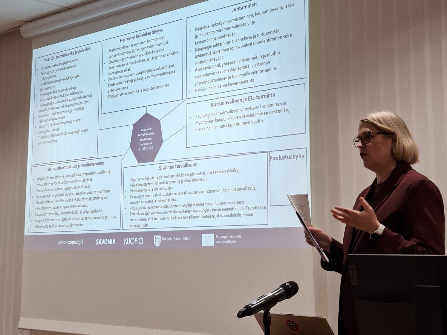 Blazeriin pukeutunut nainen seisoo puhujakorokkeen vieressä ja puhuu konferenssihuoneessa suomenkielisen tekstin ja kaavioiden täyttämän diaesityksen edessä. Pöydällä näkyy mikrofoni ja kannettava tietokone.