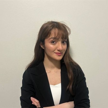 Nainen, jolla on pitkät ruskeat hiukset ja otsatukka ja jolla on musta bleiseri valkoisen topin päällä, seisoo vaalean seinän edessä kädet ristissä ja hymyilee hieman.