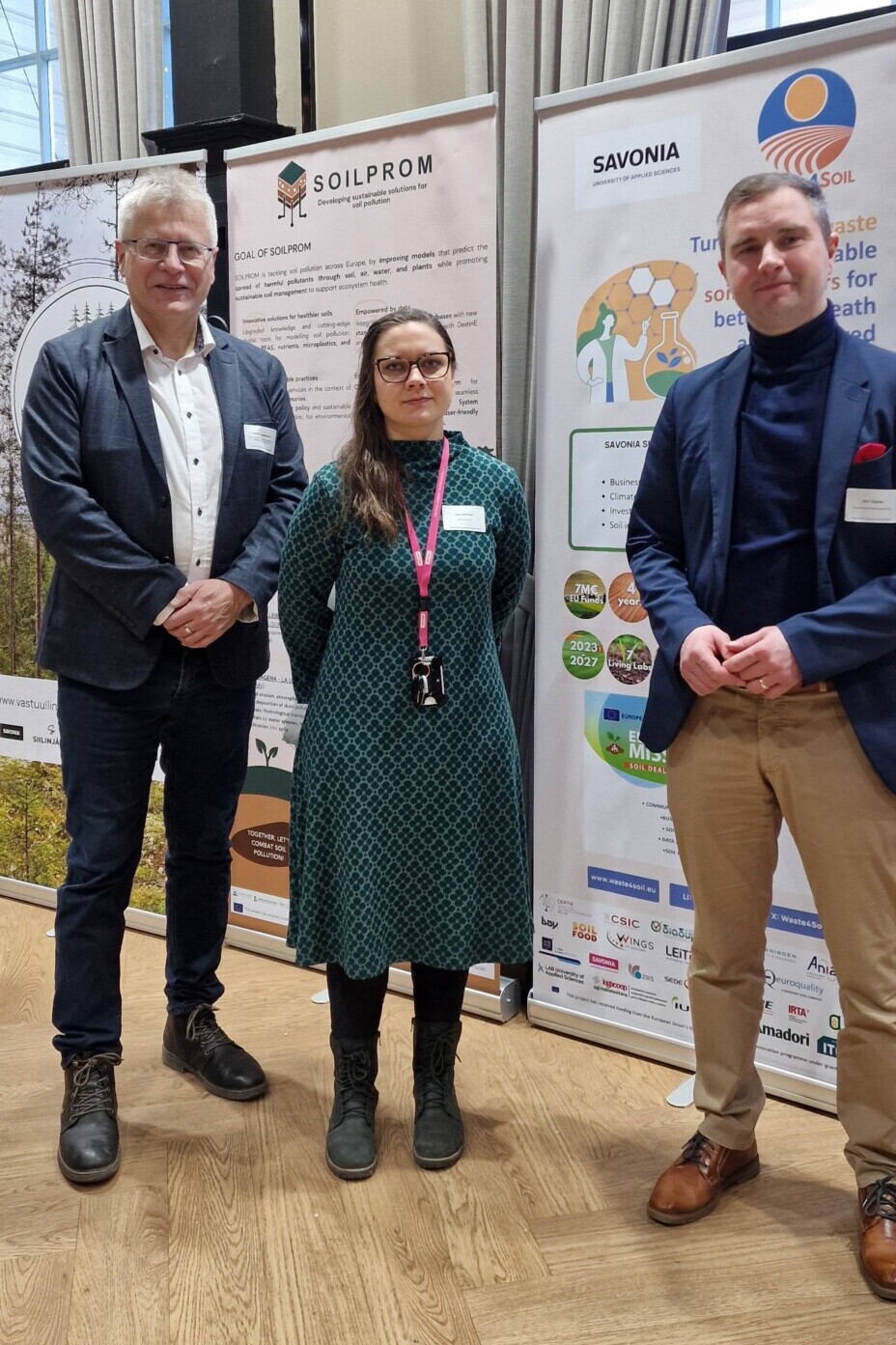 Kolme ihmistä seisoo sisätiloissa SOILPROM- ja TURF SOIL -hankkeista kertovien tiedotusbanderollien edessä. He ovat pukeutuneet rentoon työasuun ja hymyilevät kameralle. Suurista ikkunoista tuleva valo valaisee tilaa.