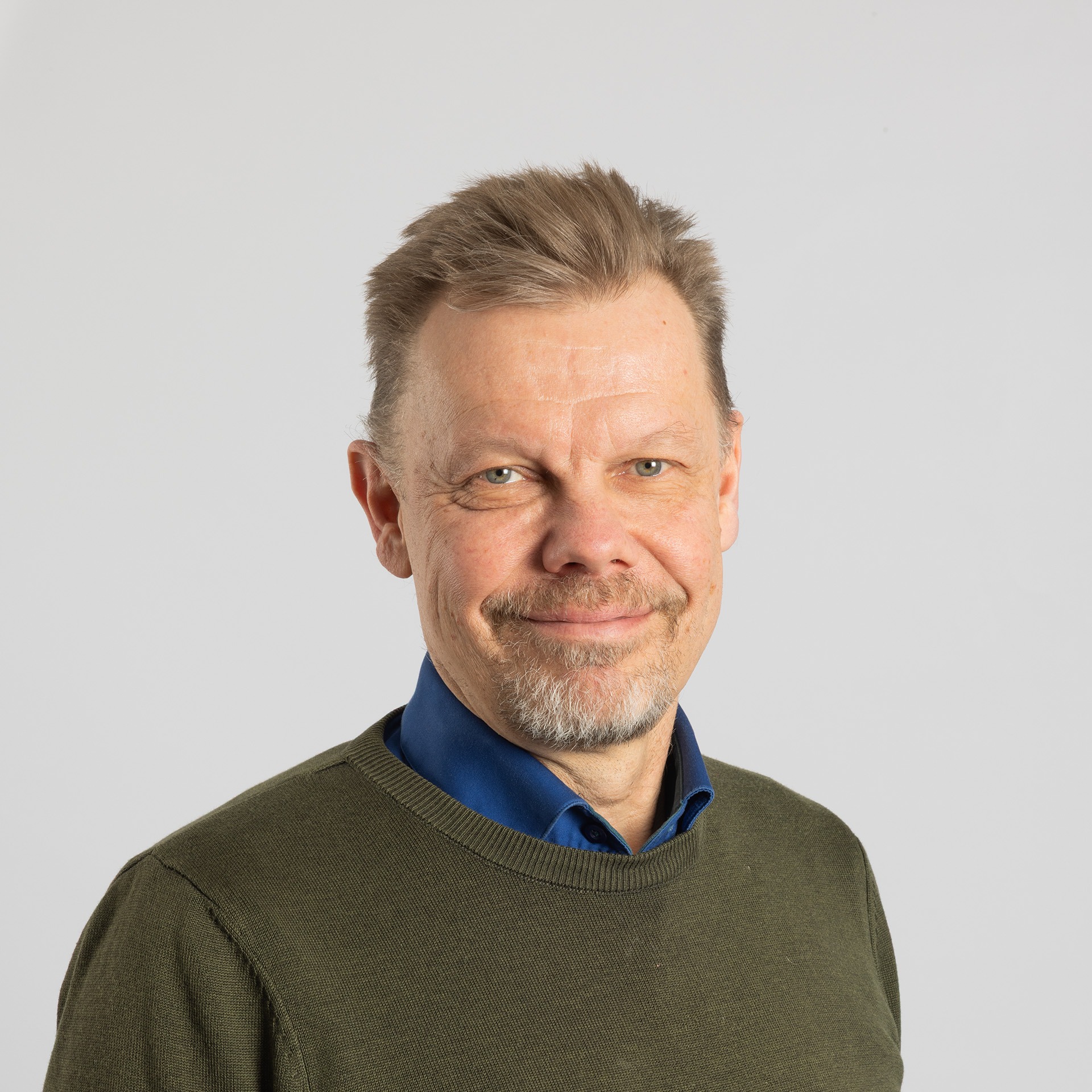 Keski-ikäinen mies, jolla on lyhyet vaaleanruskeat hiukset, vaalea parta ja viikset, hymyilee hieman. Hänellä on yllään oliivinvihreä villapaita sinisen kauluspaidan päällä, ja hän seisoo tavallisen vaaleanharmaan taustan edessä.