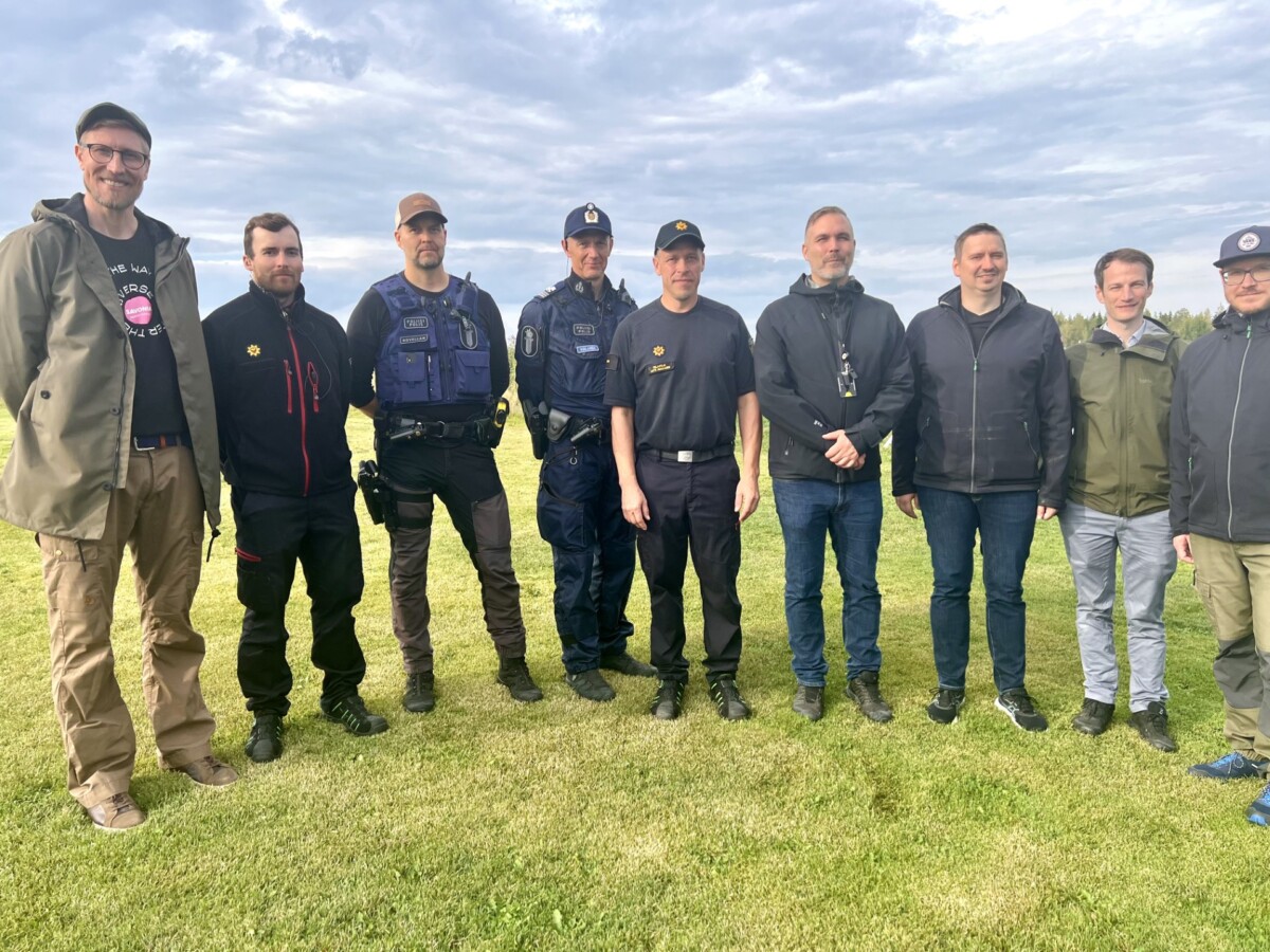 Yhdeksän hengen ryhmä, joista kahdella on poliisin univormu ja muut ulkoiluvaatteet, seisoo yhdessä ruohikolla pilvisen taivaan alla poseeraten valokuvaa varten.