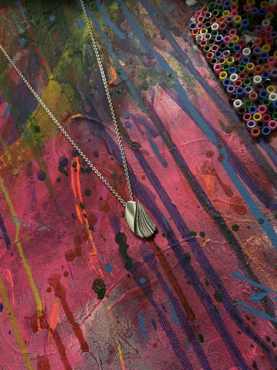 Hopeinen simpukkariipus kaulakoru makaa eloisalla, monivärisellä maalatulla pinnalla, jonka oikeassa yläkulmassa on säiliö pieniä, värikkäitä muovihelmiä.