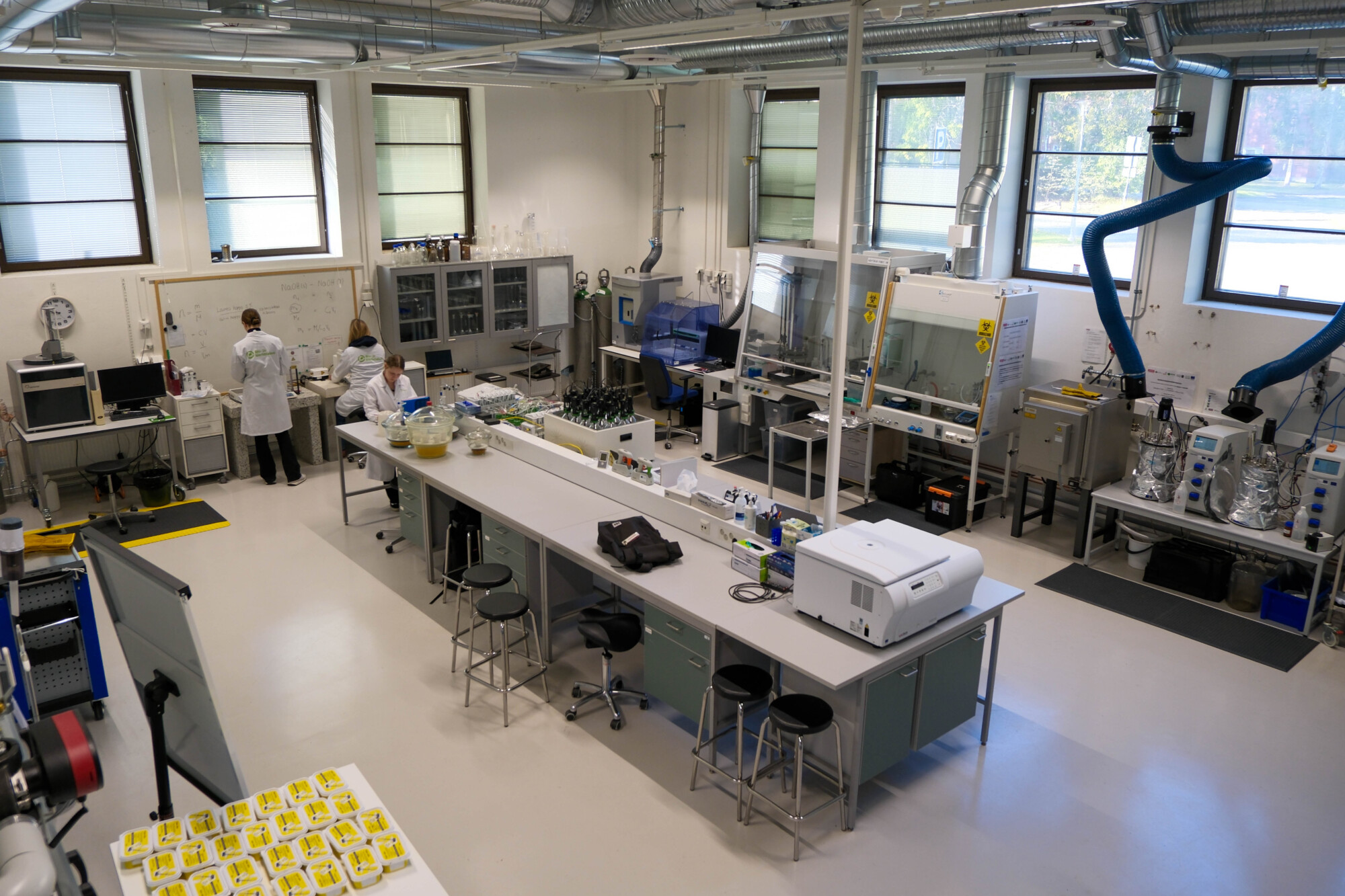 Nykyaikainen laboratorio, jossa työskentelee kaksi laboratoriotakkiin pukeutunutta ihmistä tieteellisten laitteiden, tietokoneiden, huurujen ja työpöytien ympäröimänä, ja suuret ikkunat päästävät luonnonvaloa sisään.