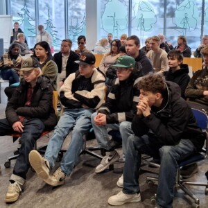 Ryhmä nuoria, useimmiten teini-ikäisiä, istuu tiiviisti yhdessä tuoleilla luokkahuoneessa tai kokoushuoneessa ja katselee tarkkaavaisesti jotakin edessä olevaa asiaa. Taustalla ovat suuret ikkunat, joissa on luonnoksia ja luminen näkymä.