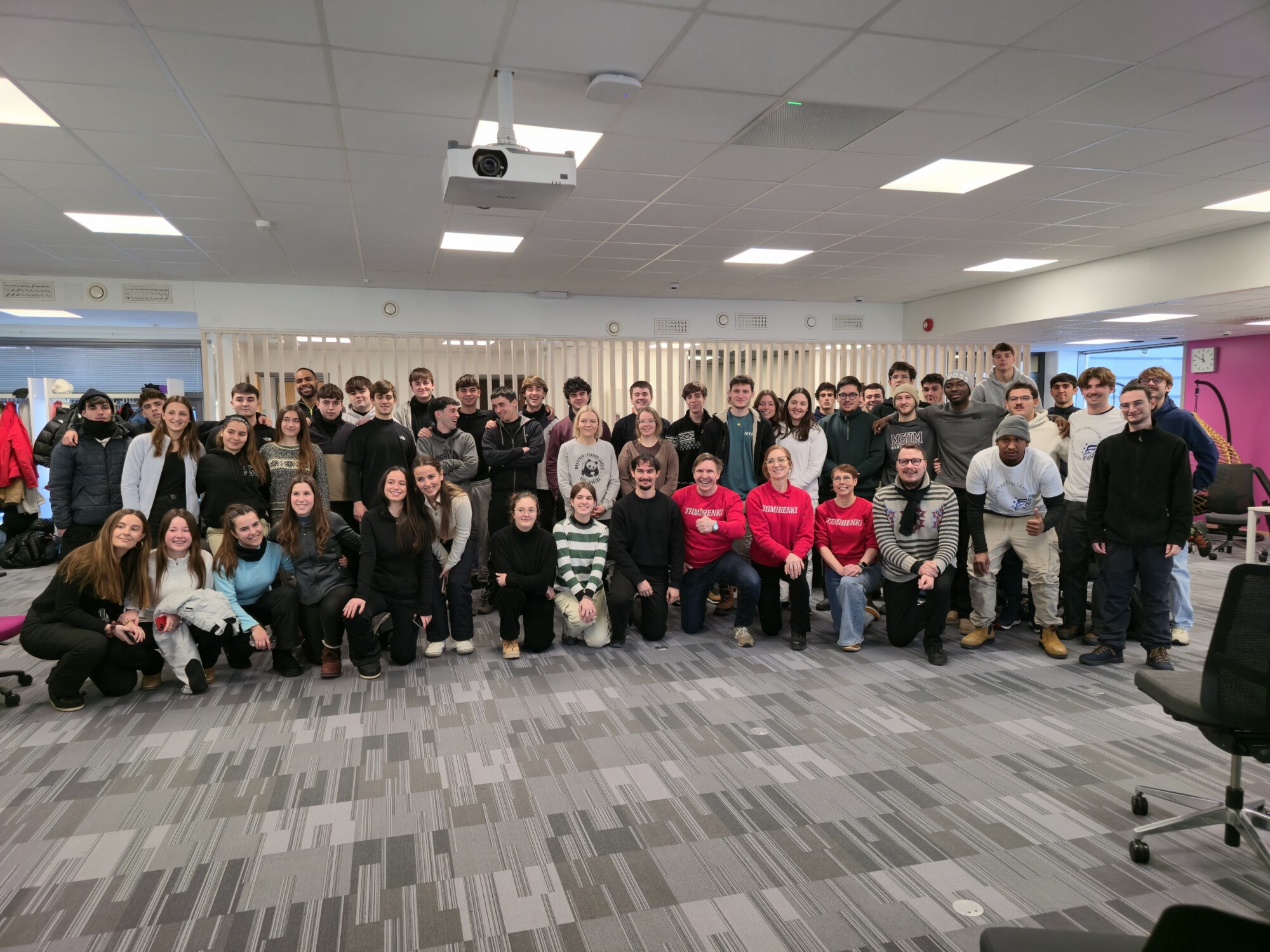 Suuri joukko ihmisiä, enimmäkseen nuoria aikuisia, poseeraa yhdessä luokkahuoneessa, jossa on harmaa matto, toimistotuoleja ja valkoiset kattolaatat. Jotkut seisovat, kun taas toiset polvistuvat tai istuvat edessä.