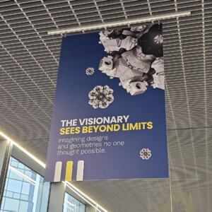 Katosta roikkuu sininen banderolli, jossa on geometrisia 3D-muotoja ja teksti: NÄKIJÄ NÄKEE YLI RAJOJEN. Kuvittelee malleja ja geometrioita, joita kukaan ei pitänyt mahdollisina. Keltaiset ja valkoiset korostukset korostavat kylttiä.