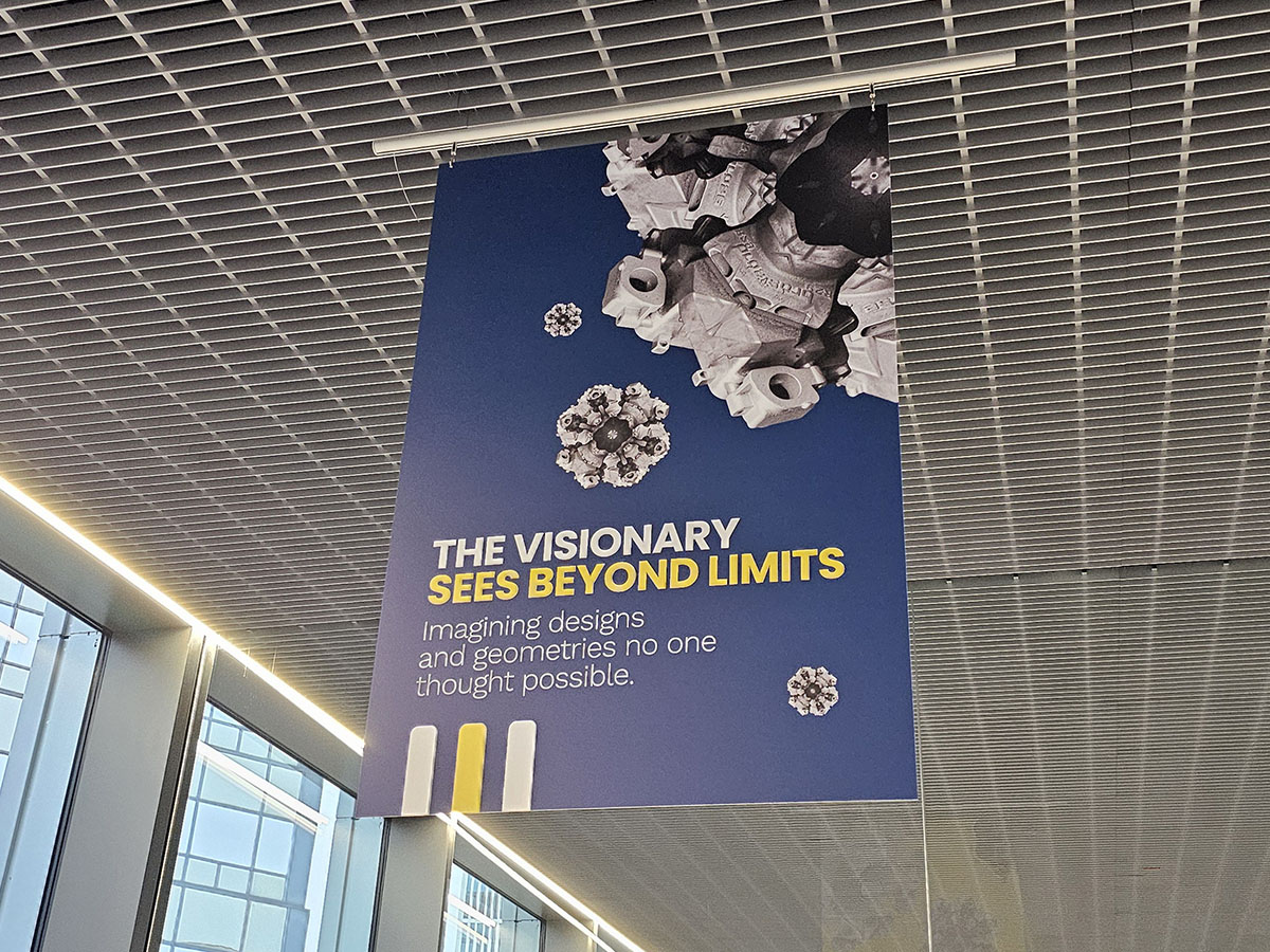 Katosta roikkuu sininen banderolli, jossa on geometrisia 3D-muotoja ja teksti: NÄKIJÄ NÄKEE YLI RAJOJEN. Kuvittelee malleja ja geometrioita, joita kukaan ei pitänyt mahdollisina. Keltaiset ja valkoiset korostukset korostavat kylttiä.