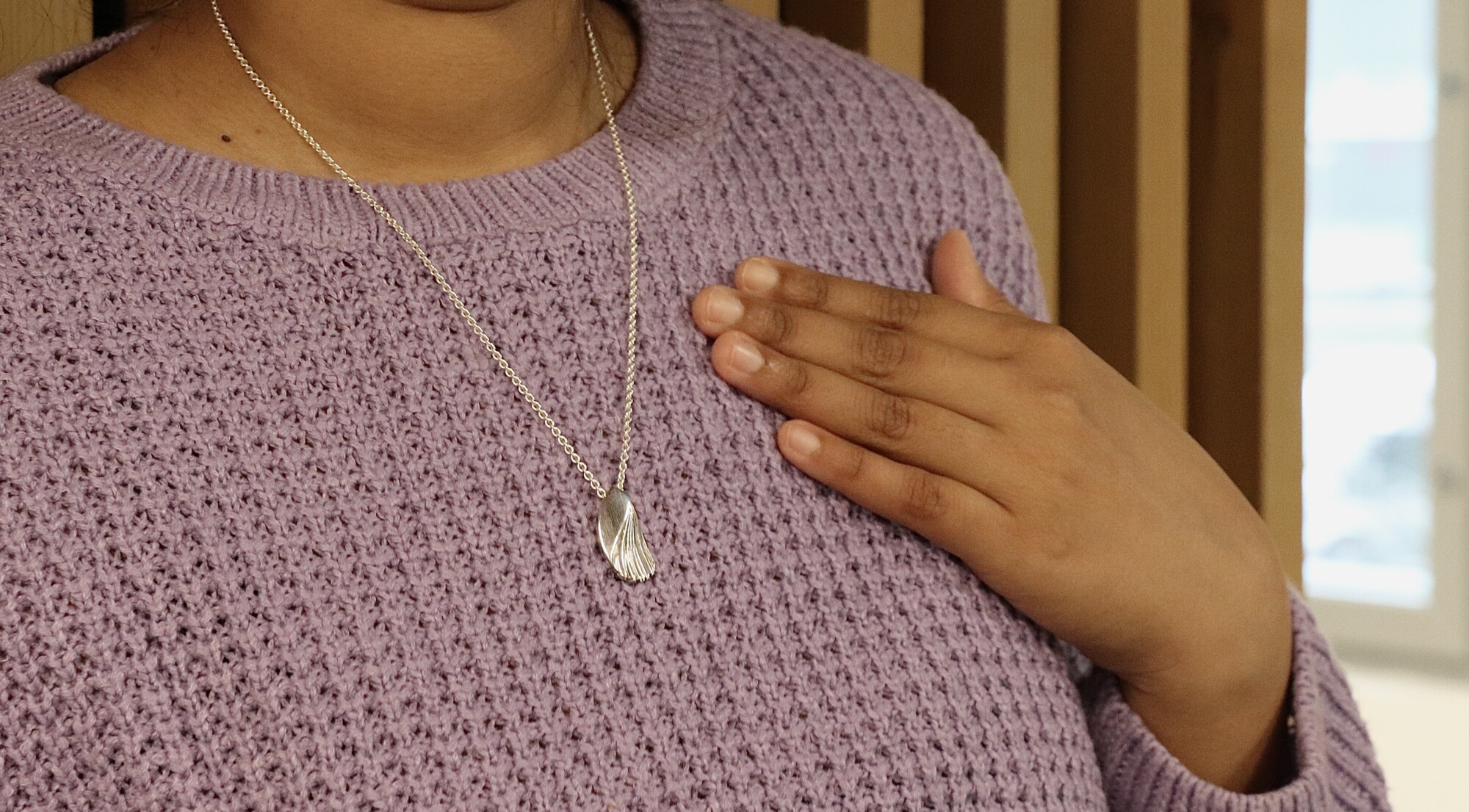 Henkilö, jolla on yllään kuvioitu violetti villapaita, koskettaa toisella kädellään rintaansa, jossa on hopeinen riipuskaulakoru. Taustalla on pystysuuntaisia puisia säleikköjä ja hämärtynyt ikkuna.