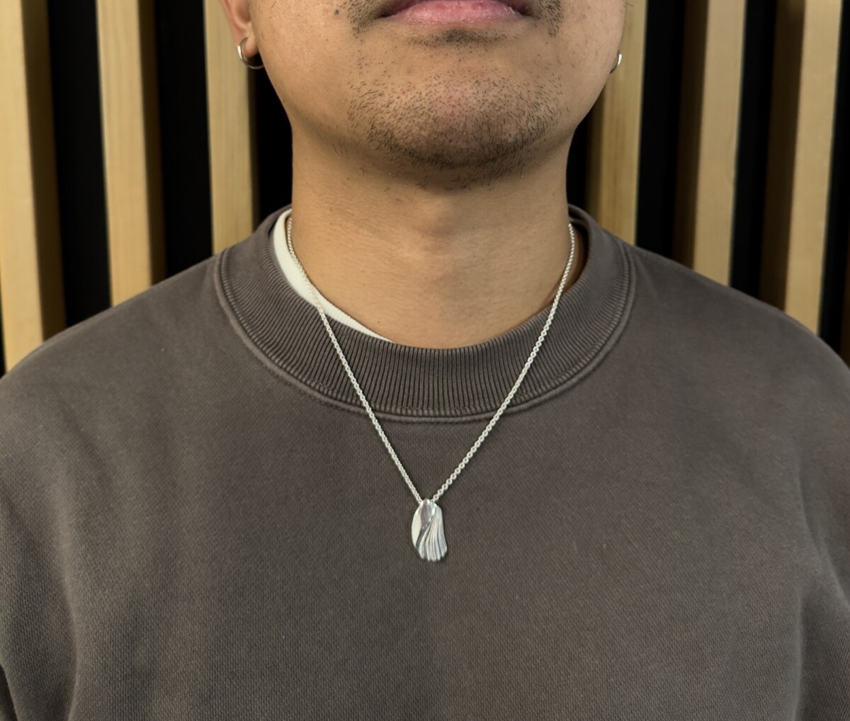 Henkilö, jolla on tummanruskea kauluspaita ja hopeinen kaulakoru, jossa on taitettujen siipien muotoinen riipus, seisoo puisen säleikköseinän edessä.