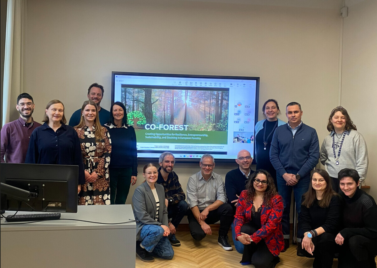 Viidentoista ihmisen ryhmä poseeraa ja hymyilee luokkahuoneessa, seisoo ja polvistuu suurella näytöllä olevan esitysdian edessä, jonka otsikkona on CO-FOREST ja jossa on metsäkuva ja teksti Euroopan metsistä.