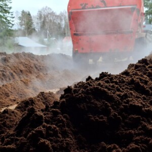 Suurella punaisella maatalouskoneella käännetään tai käsitellään ulkona tummia maakasoja, ja taustalla näkyy puita ja rakennuksia.