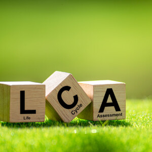 Vihreällä ruoholla on kolme puupalikkaa, joissa lukee LCA, ja niiden alla on pienempi teksti, jossa lukee Life Cycle Assessment (elinkaariarviointi). Taustalla on kirkas, hämärtynyt vihreä.