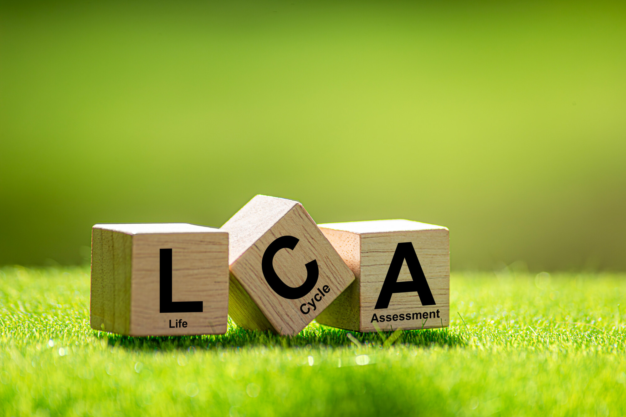 Vihreällä ruoholla on kolme puupalikkaa, joissa lukee LCA, ja niiden alla on pienempi teksti, jossa lukee Life Cycle Assessment (elinkaariarviointi). Taustalla on kirkas, hämärtynyt vihreä.