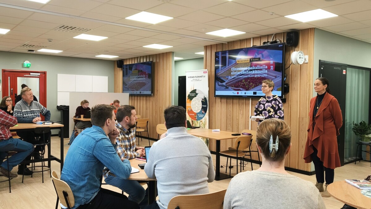 Joukko ihmisiä istuu pöydissä valoisassa luokkahuoneessa ja kuuntelee kahta naista, jotka seisovat ja esittelevät suurella näytöllä, jolla näkyy värikäs grafiikka ja teksti.