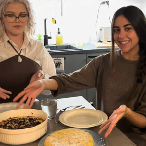 Kaksi naista istuu keittiön pöydän ääressä, hymyilee ja esittelee ruokalajeja edessään - toinen on paistettu vuoka, toinen pyöreä litteä leipä. Keittiössä on luonnonvaloa ja se näyttää siistiltä.