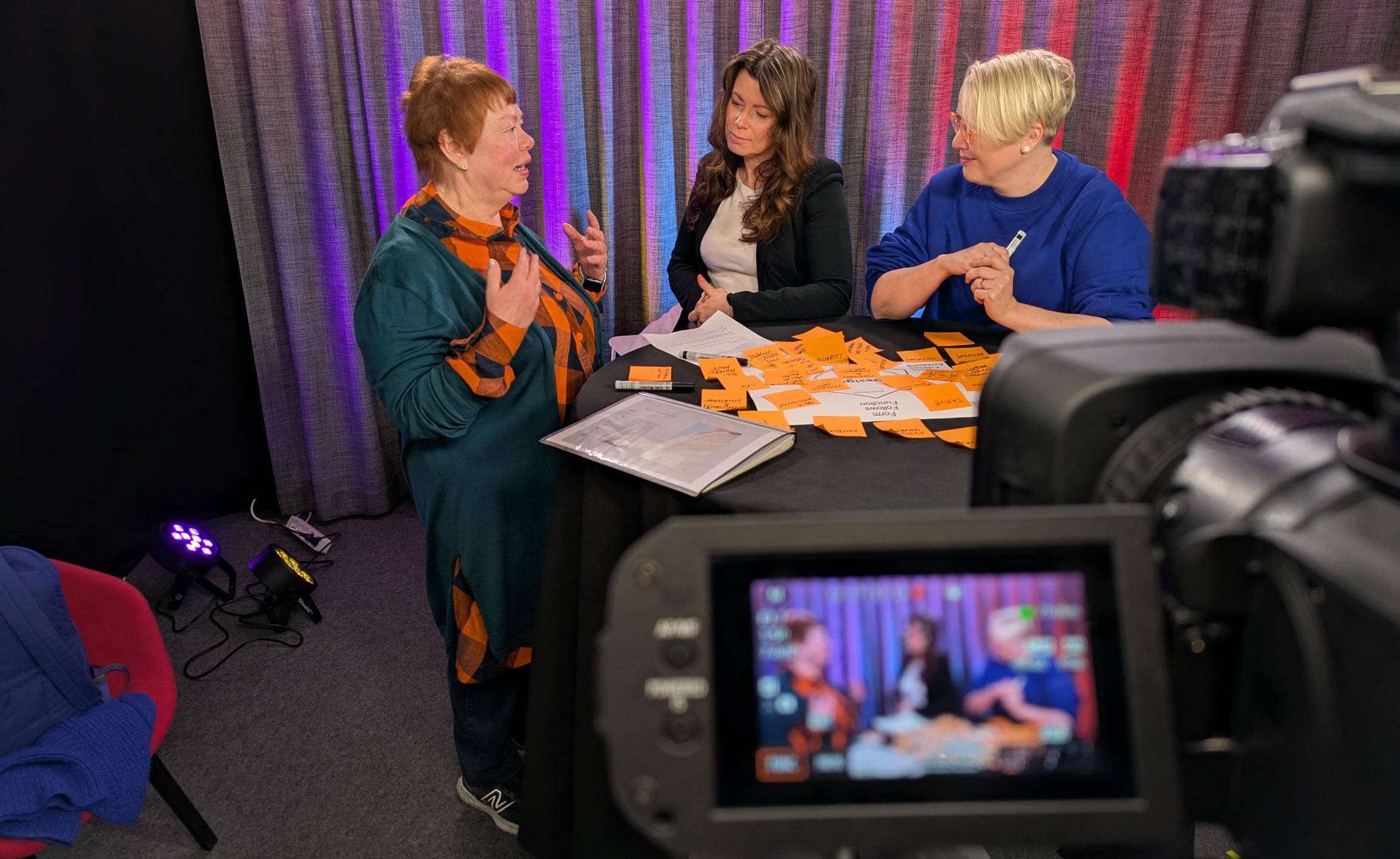 Kolme naista istuu oransseilla muistilapuilla päällystetyn pöydän ympärillä keskustelemassa, ja etualalla oleva kamera kuvaa keskustelua. Taustalla on violetilla valolla valaistu verho.