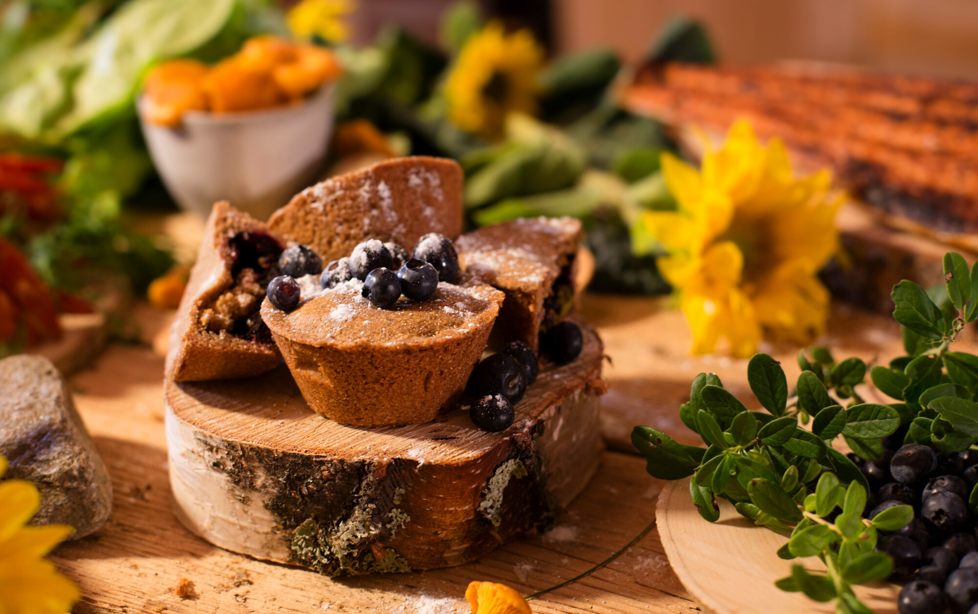 Lähikuva pienistä, pyöreistä, mustikoilla ja tomusokerilla päällystetyistä kakkuista, jotka ovat esillä maalaismaisella puupinnalla vihreiden lehtien, keltaisten kukkien ja erilaisten ruokien sumean taustan ympäröimänä.