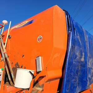 Suuri oranssi teollisuuskone on osittain peitetty sinisellä pressulla. Koneen kylkeen on kiinnitetty useita metallitankoja ja valkoinen säiliö, ja taustalla on kirkkaan sininen taivas.