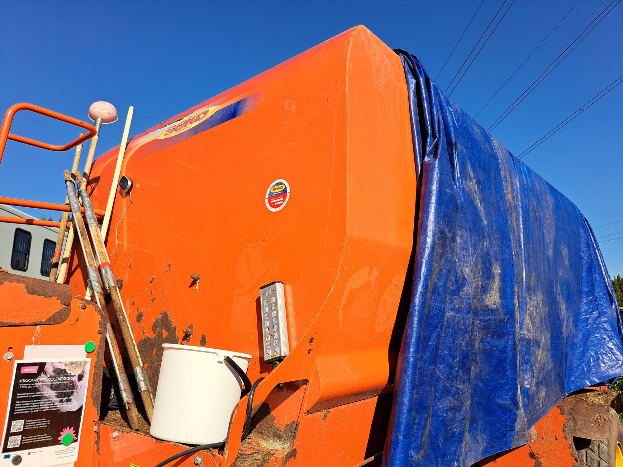 Suuri oranssi teollisuuskone on osittain peitetty sinisellä pressulla. Koneen kylkeen on kiinnitetty useita metallitankoja ja valkoinen säiliö, ja taustalla on kirkkaan sininen taivas.