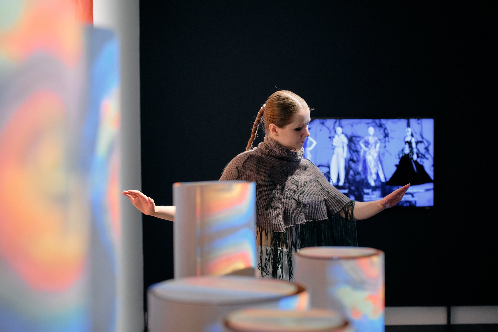 Nainen, jolla on punotut hiukset ja hapsullinen huivi, ojentaa kätensä värikkäiden, hämärtyneiden lieriömäisten esineiden keskellä. Taustalla mustavalkoisella valkokankaalla näkyy ihmisiä dramaattisissa puvuissa.