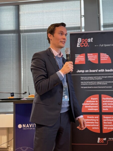 Pukumies pitelee mikrofonia ja puhuu sisätiloissa järjestettävässä tapahtumassa. Hänen takanaan on bannereita, joissa on tekstiä Boost ECOsta ja innovaatiosta. Suuret ikkunat päästävät luonnonvaloa sisään.