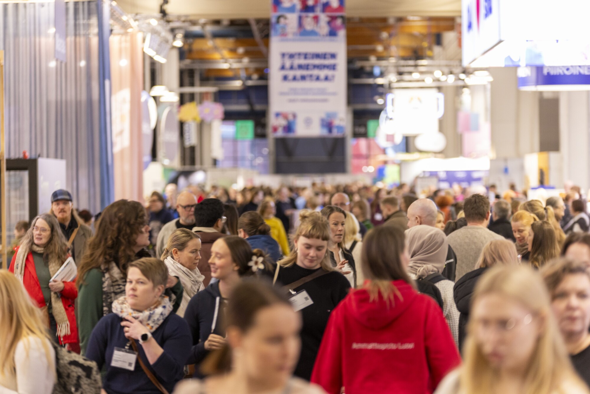 Suuri ihmisjoukko kävelee kiireisen sisätiloissa järjestettävän tapahtuman tai näyttelyhallin läpi, ja taustalla näkyy värikkäitä banderolleja ja kojuja. Tunnelma vaikuttaa vilkkaalta ja vilkkaalta.