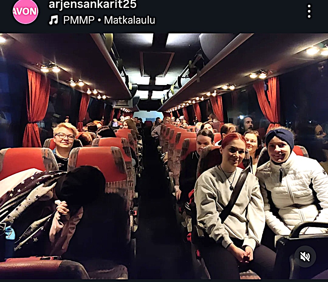 Joukko ihmisiä istuu linja-autossa, jossa on punaiset verhot, ja jotkut hymyilevät kameralle. Bussi on enimmäkseen täynnä ja hämärästi valaistu. Useilla matkustajilla on reput tai laukut.