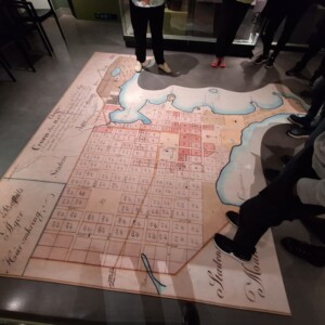 Joukko ihmisiä seisoo lattialle asetetun suuren historiallisen kaupunkikartan ympärillä, jossa on numeroitujen tonttien, vesistöjen ja ranskankielisten käsinkirjoitettujen merkintöjen ruudukko.