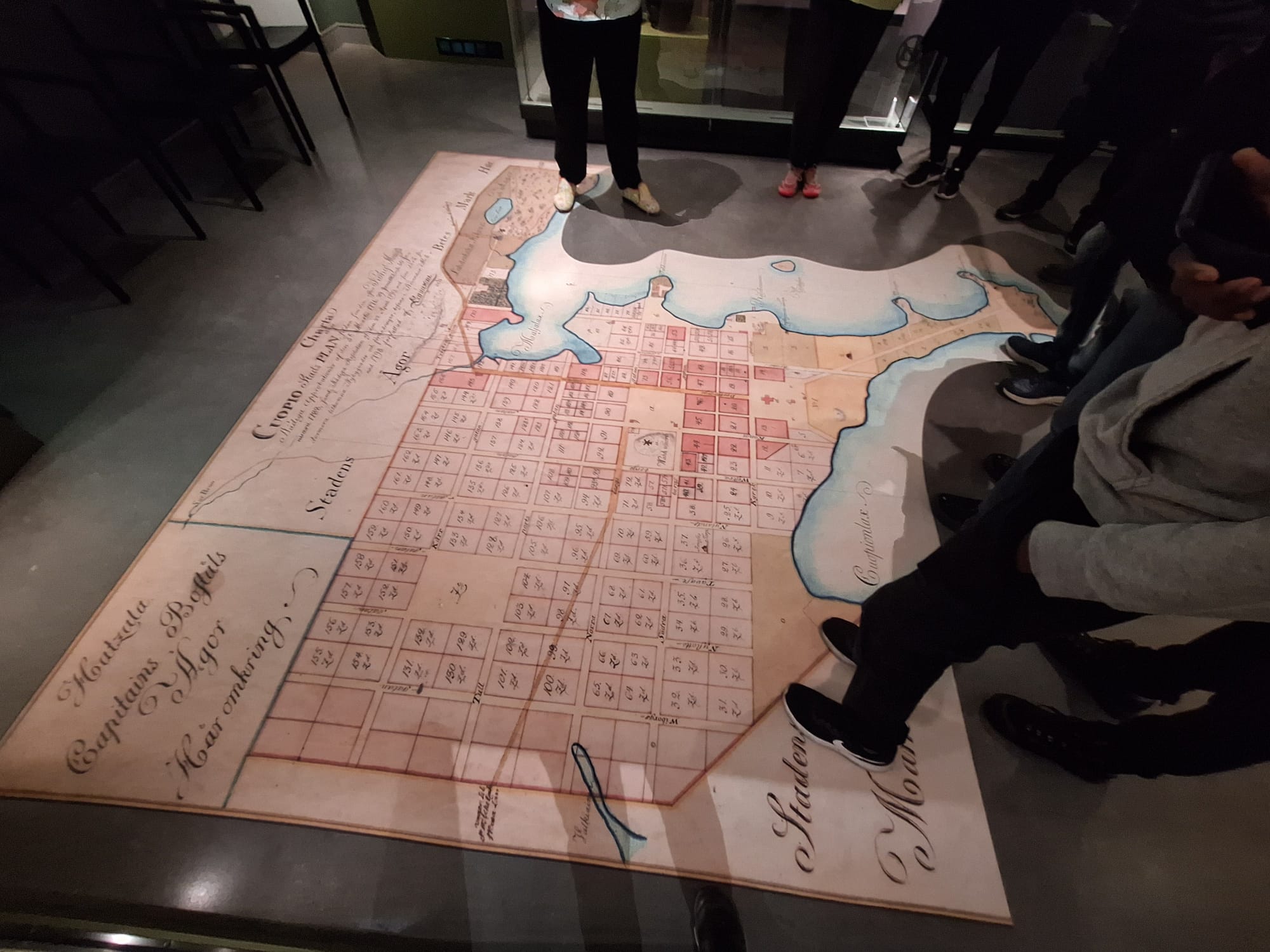 Joukko ihmisiä seisoo lattialle asetetun suuren historiallisen kaupunkikartan ympärillä, jossa on numeroitujen tonttien, vesistöjen ja ranskankielisten käsinkirjoitettujen merkintöjen ruudukko.