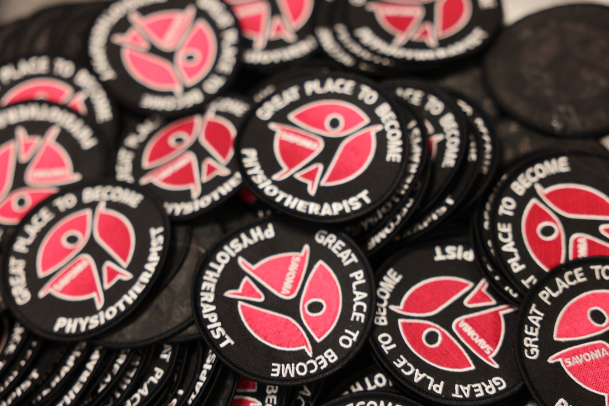 Kasa pyöreitä mustia laastareita, joissa on punavalkoinen abstrakti kuvio ja reunalla valkoisin kirjaimin teksti GREAT PLACE TO BECOME PHYSIOTHERAPIST.