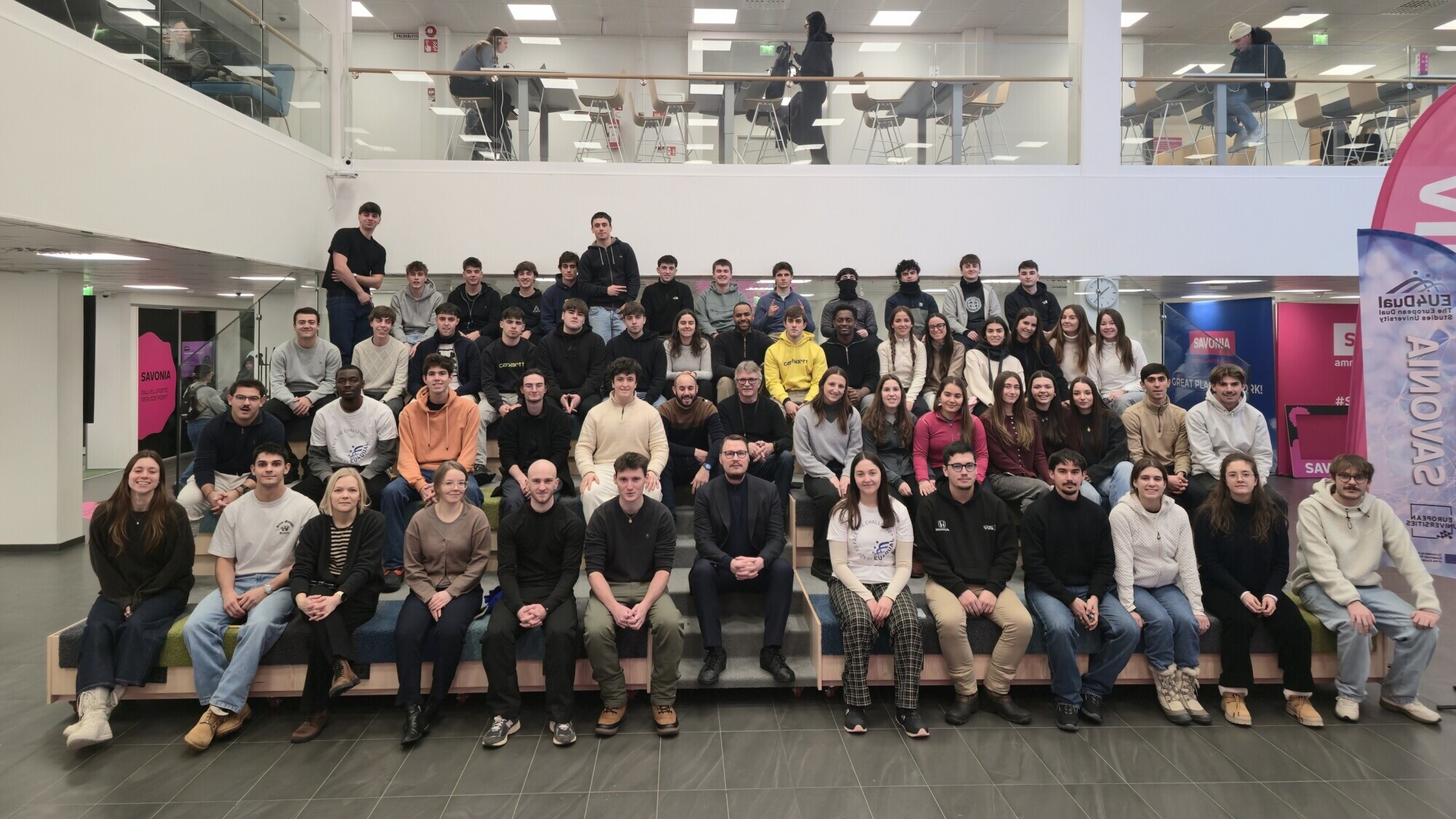 Suuri joukko ihmisiä, enimmäkseen nuoria aikuisia, istuu ja seisoo porrastetuilla portailla ryhmäkuvaa varten modernissa sisätilassa, jossa on lasikaiteet ja yläpuolella työskentelevät ihmiset.