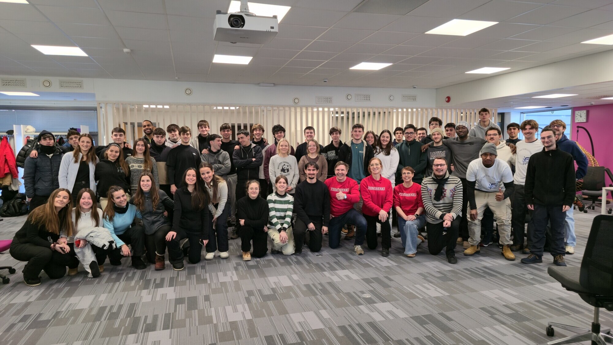 Suuri joukko ihmisiä, enimmäkseen nuoria aikuisia, poseeraa ja hymyilee yhdessä kirkkaasti valaistussa huoneessa, jossa on harmaa matto, valkoiset kattolaatat ja toimistotuolit. Jotkut polvistuvat edessä, kun taas toiset seisovat heidän takanaan.